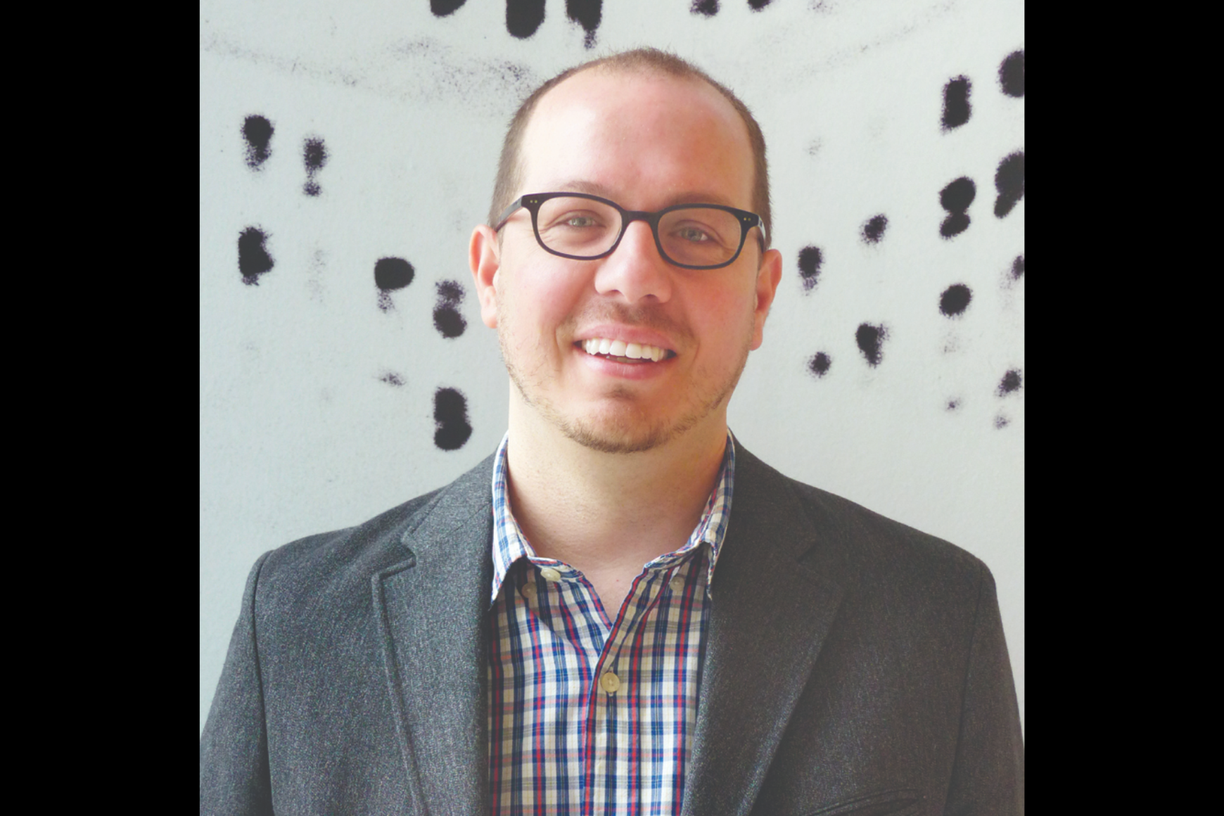 Man wearing glasses and a grey suit smiling in front of a black and white wall